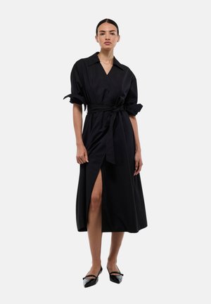 Robe noire avec des manches courtes retroussées, une ceinture à la taille et une fente sur le côté. Tissu lisse avec un éclat subtil, associée à des chaussures noires à talons pointus.