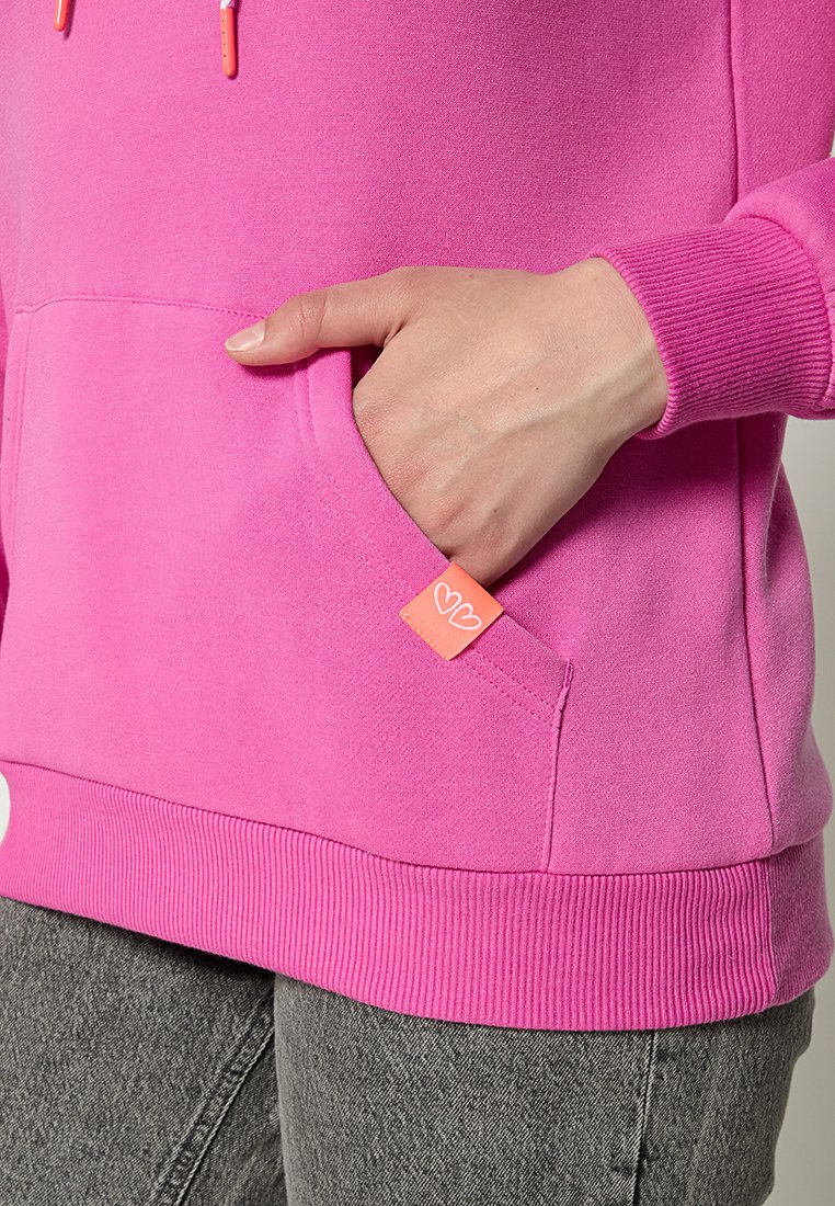 Fuchsia Hoodie aus weichem Stoff, mit einer Kängurutasche und einem kleinen orangen Etikett. Gerippte Bündchen und Saum sorgen für zusätzliche Textur.