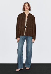 Model stands confidently in a brown suede overshirt and beige knit top, paired with wide-leg blue jeans and black sandals against a light backdrop.