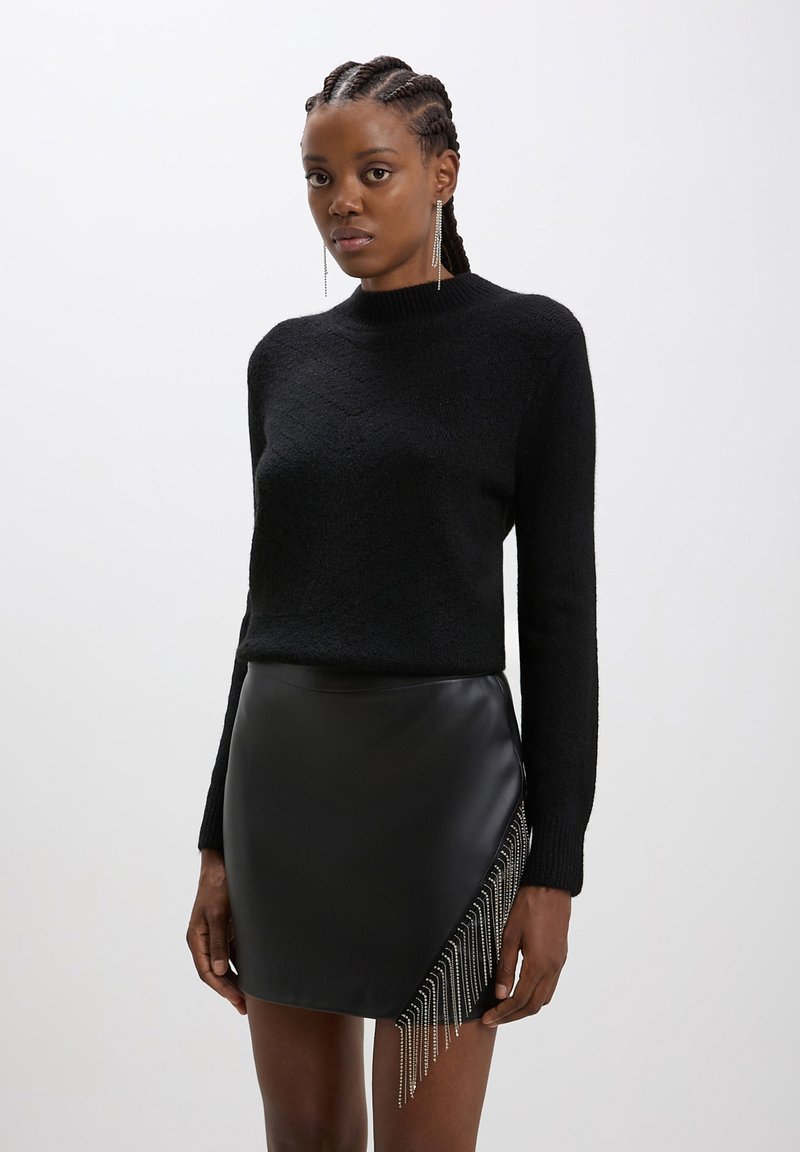 Donna che indossa un maglione lungo a maniche nere con texture e una mini gonna di pelle nera con dettagli in frange argentate, in piedi contro uno sfondo chiaro uniforme.