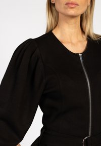 Zwarte zip-up jas met opgezette mouwen, gemaakt van gestructureerde stof. Bevat een zilveren rits en een stoffen riem met een ronde gesp.