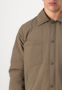 Khaki button-up jacket made of lightweight fabric, featuring a collar, front pocket, and visible stitching.