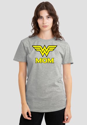 Woman with brown hair wears gray T-shirt with yellow Wonder Woman logo and word "MOM" in bold letters on a plain background.