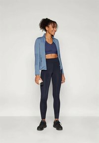 Vaaleansininen vetoketjupusakka tummansinisellä urheilurintaliivillä, yhdistettynä korkeavyötäröisiin tummiin legginsseihin. Mustat urheilukengät ja käsipaino kädessä.