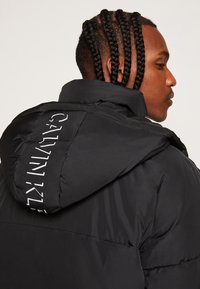 Giacca a vento nera con una texture liscia, dotata di cappuccio e logo Calvin Klein bianco sul retro. Design strutturato con un taglio moderno.