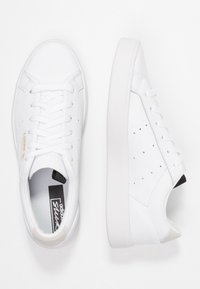 Vita sneakers med en slät läderövre, platta snören, minimala perforeringar, grå accenter på hälen och en vit gummisula.