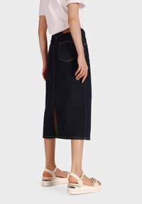 Gonna midi in denim scuro con spacco laterale, tasche posteriori e cuciture a contrasto. Abbinata a sandali beige e a una blusa leggera.