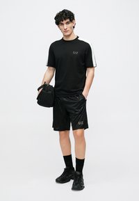 Schwarzes T-Shirt mit weißen Schulterstreifen, kombiniert mit schwarzen Shorts mit geometrischem Muster und goldenem Logo. Accessoire: kleine schwarze Tasche.