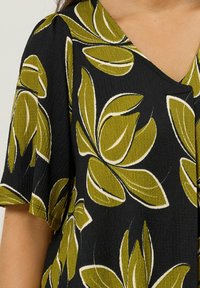 Tissu noir avec une finition texturée, présentant de grands motifs floraux verts et blancs et un décolleté en V. Des manches courtes mettent en valeur le design.