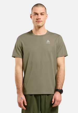 Man standing facing forward wearing olive green short-sleeve t-shirt and matching pants, with a watch on left wrist.