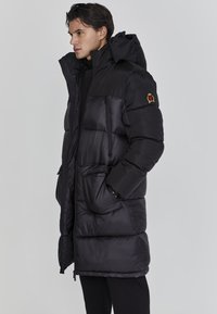 Mustavi musta takki, jossa on huppu, tikatut osiot, etuvetoketju, sivutaskut ja emblem vasemmassa hihassa.