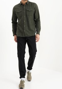 Olivgrön flanellskjorta med två bröstfickor, knäppt med en texturerad yta, kombinerad med svarta byxor och beige sneakers.