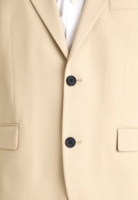 Beige blazer lavet af tekstureret stof med to sorte knapper, slåede kraver og to forlommer.