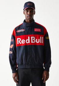 Μπουφάν εμπνευσμένο από την ομάδα Red Bull Racing σε ναυτικό μπλε και κόκκινο, με λογότυπα, ψηλό γιακά, ριμπ μανσέτες και φερμουάρ μπροστά. Το καπέλο συμπληρώνει την εμφάνιση.