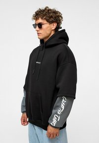 Zwarte oversized hoodie met een voorzak en een verstelbare trekkoordkap. Grijze mouwen met de tekst "Creative Care" in het wit. Zachte stoftextuur.
