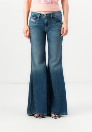 Persona vestida con jeans de mezclilla oscuros de tiro alto y las sandalias de tacón abierto, de pie frente a un fondo blanco plano.