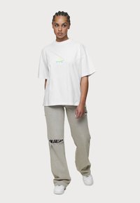 Weißes oversized T-Shirt mit Druckgrafiken auf der Brust, kombiniert mit hellgrauen, distressed Jeans mit Rissen an den Knien und weißen Sneakers.