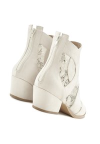 Botas de tobillo de ante sintético blanco con tacón grueso. Cuenta con recortes en forma de cordones y un cierre de cremallera lateral para facilitar su uso. Textura suave, diseño moderno.