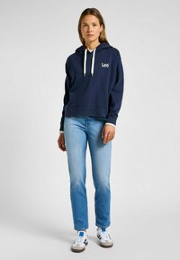 Sudadera de color azul marino con un diseño corto, cordones blancos y logo bordado. Combinada con jeans de color azul claro y zapatillas casuales.