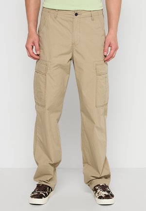 Uomo che indossa pantaloni cargo beige con tasche laterali e sneakers nere e bianche con motivo, in piedi davanti a uno sfondo semplice.