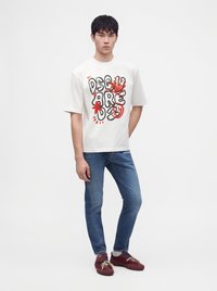 Jeune homme portant un t-shirt blanc à motif graphique, un jean bleu et des baskets rouges, debout devant un fond uni clair.