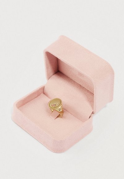 Kultasormus, jonka yläosa on soikea ja koristeltu kivillä, asetettuna pehmeään vaaleanpunaiseen samettiseen sormusrasiaan. Rasian sisäosa vastaa ulkopuolen väriä ja rakennetta.