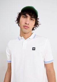 Hvid polo-shirt med blå-stribede krave og manchetter, prydet med et lille sort logo-patch. En grøn cap komplementerer outfittet.
