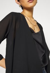 Blouse noire transparente avec des manches drapées superposée à un camisole en satin noir. Les bijoux comprennent des chaînes en or superposées et des boucles d'oreilles créoles.