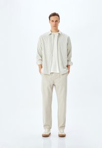 Gestreifte, helle Button-Up-Bluse über einem weißen T-Shirt, kombiniert mit beigen, weit geschnittenen Hosen und hellen Sneakers mit brauner Sohle.
