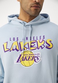 Svetlo modra kapuca z izrazitim vijoličnim in rumenim besedilom "Los Angeles Lakers" ter grafiko košarke, ki je središča na prsih. Bombažni material.
