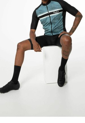 Uomo seduto su un cubo bianco che indossa scarpe da ciclismo nere, calze nere, maglia da ciclismo nero e blu e pantaloncini da ciclismo neri.