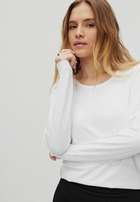 Langärmliges weißes Oberteil aus glattem Stoff mit Rundhalsausschnitt. Das Modell hat die Hände unter dem Kinn verschränkt und präsentiert ein minimalistisches Design.