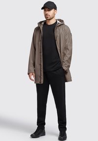 Taupe vandtæt jakke med hætte, sort t-shirt og sorte sweatpants. Udstyret med trykknapper og båret med sorte sneakers.