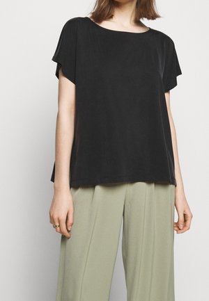 Person wearing a loose black short-sleeve top and light green wide-leg pants, standing against a plain light background.