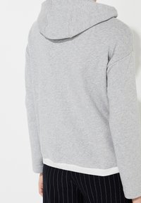 Sudadera con capucha de forro polar gris, con un ribete blanco en la parte inferior. La tela es suave y ligeramente texturizada, con un ajuste holgado.