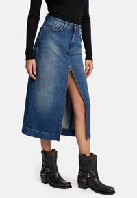 Jupe maxi en denim avec une fermeture éclair à l'avant et une fente ; présente un lavage bleu foncé et une silhouette droite. Associée à des bottines noires.