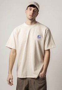 Młody mężczyzna w beżowej, oversize'owej koszulce z małym niebieskim grafiką, w pasującej beżowej czapce i brązowych spodniach, stoi na jasnym, jednolitym tle.