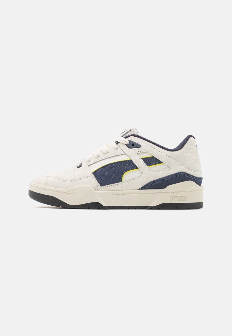 Vue latérale d'une basket Puma blanche et bleu marine avec fermeture à lacets et semelle noire sur fond uni.