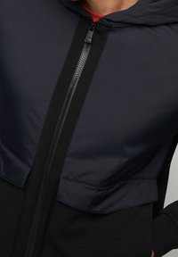 Giacca con cappuccio caratterizzata da una parte superiore di un blu navy scuro con una texture liscia e una parte inferiore nera. Chiusura lampo frontale e due tasche grandi.