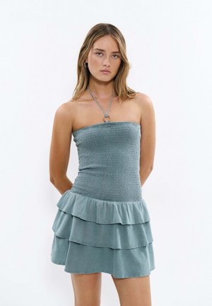 Junge Frau trägt ein trägerloses, hellblaues Kleid mit gerafftem Oberteil und mehrlagigem Rüschenrock, steht mit den Händen hinter dem Rücken vor einem schlichten weißen Hintergrund.