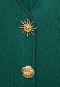 Tissu tricoté vert avec deux boutons décoratifs dorés, l'un en forme de soleil avec des rayons et l'autre avec une surface froissée et texturée.