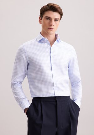 Camicia a quadri azzurri chiari con maniche lunghe, colletto button-down e tessuto leggero, abbinata a pantaloni scuri, mani nelle tasche.