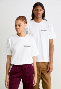 Weiße Baumwoll-T-Shirts mit kurzen Ärmeln, die ein schwarzes Textlogo auf der linken Brust tragen. Das Modell trägt dazu eine burgunderrote und braune Hose.