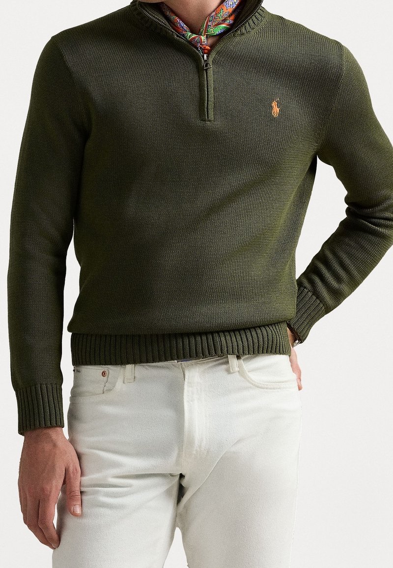Homme portant un pull vert olive à demi-fermeture éclair avec un petit logo brodé, un foulard en soie à motifs autour du cou, et un pantalon blanc, main dans la poche.