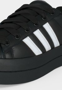 Schwarzer Ledersneaker mit schwarzen Schnürsenkeln und drei weißen Zickzack-Streifen an der Seite, Nahaufnahme.