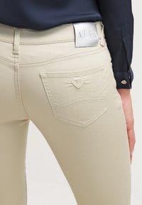 Beiga jeans med en kroppsnära design. Har en silvrig "AJ" logotyp patch vid midjan och en liten broderad logotyp på bakfickan.