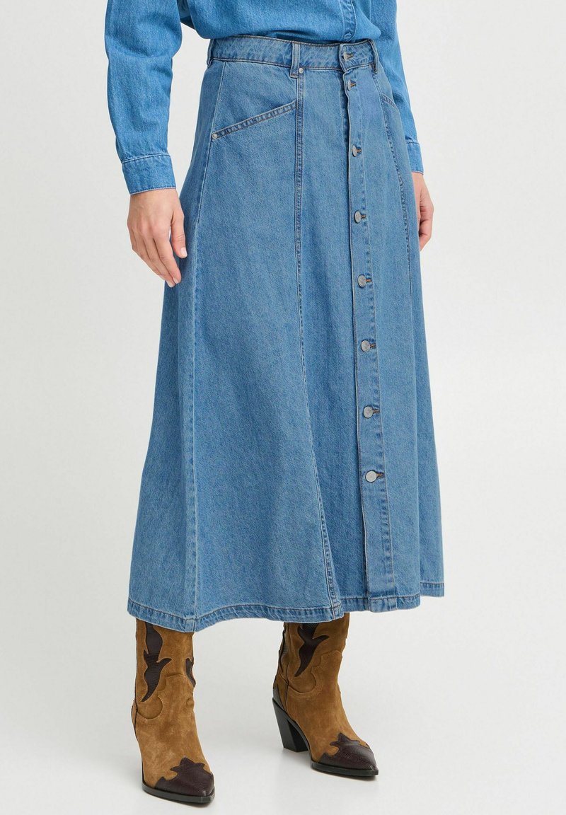 Jupe maxi en denim bleu avec fermeture à boutons sur le devant, poches latérales et design légèrement évasé. Portée avec des bottines brunes à la cheville arborant un motif.