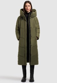 Dlhý prešívaný kabát v olivovo zelenej farbe s vysokým golierom, cvokovými gombíkmi a prešívaným vzorom, nosený cez čierny top a kombinovaný s čiernymi čižmami.