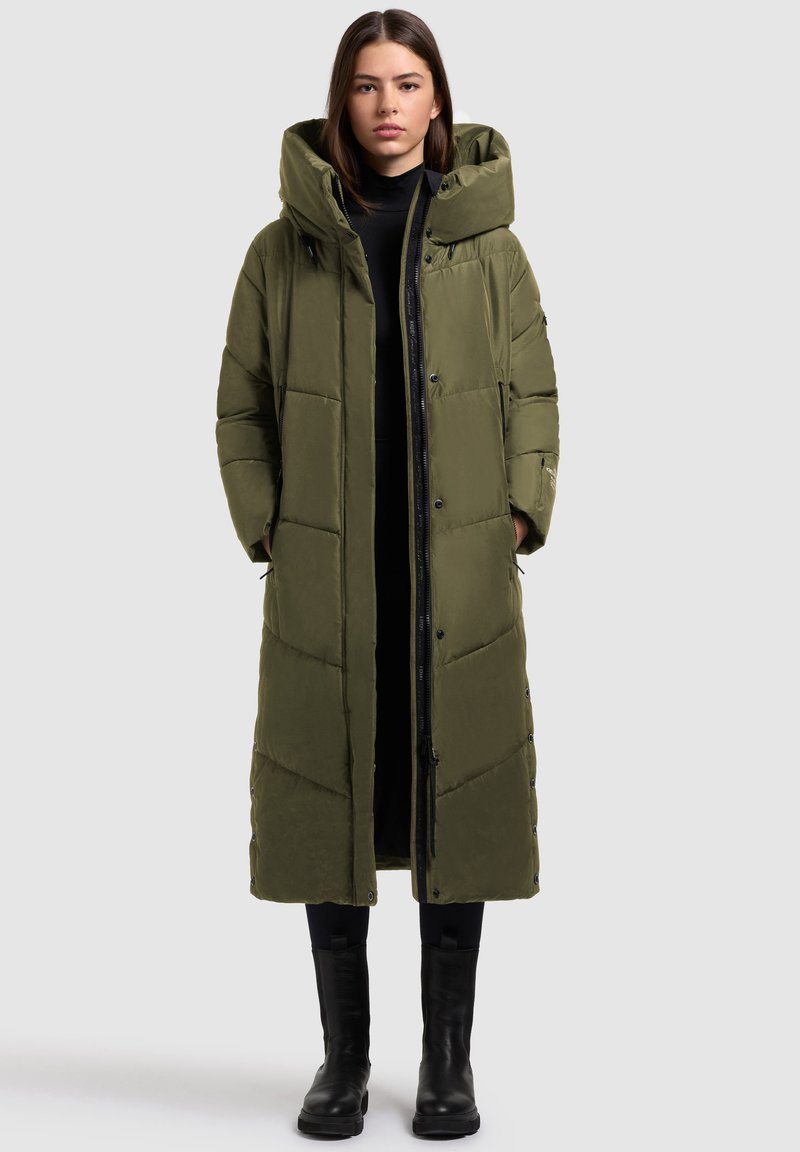 Dlhý prešívaný kabát v olivovo zelenej farbe s vysokým golierom, cvokovými gombíkmi a prešívaným vzorom, nosený cez čierny top a kombinovaný s čiernymi čižmami.