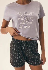 Jasnofioletowa bawełniana koszulka z białym napisem i kwiatowym wzorem, w połączeniu z czarnymi szortami z białym kwiatowym wzorem i elastycznym paskiem.